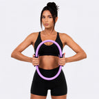 A woman in a black sports bra and shorts holds a purple exercise hoop against her torso, posed against a light gray studio background.
