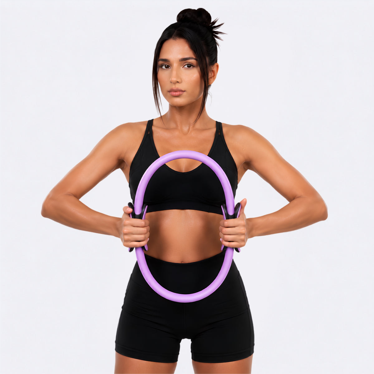 A woman in a black sports bra and shorts holds a purple exercise hoop against her torso, posed against a light gray studio background.