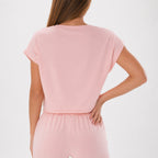 A woman shown from the back wearing a light pink short-sleeve top and matching pink bottoms against a plain light gray studio background.