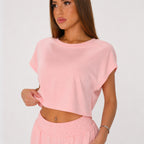 A woman wearing a light pink short-sleeve crop top and matching pink pants, photographed against a plain light gray studio background.