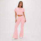 A woman modeling a light pink two-piece outfit consisting of a short-sleeve cropped t-shirt and matching high-waisted flare pants, paired with white sneakers against a plain white studio background.