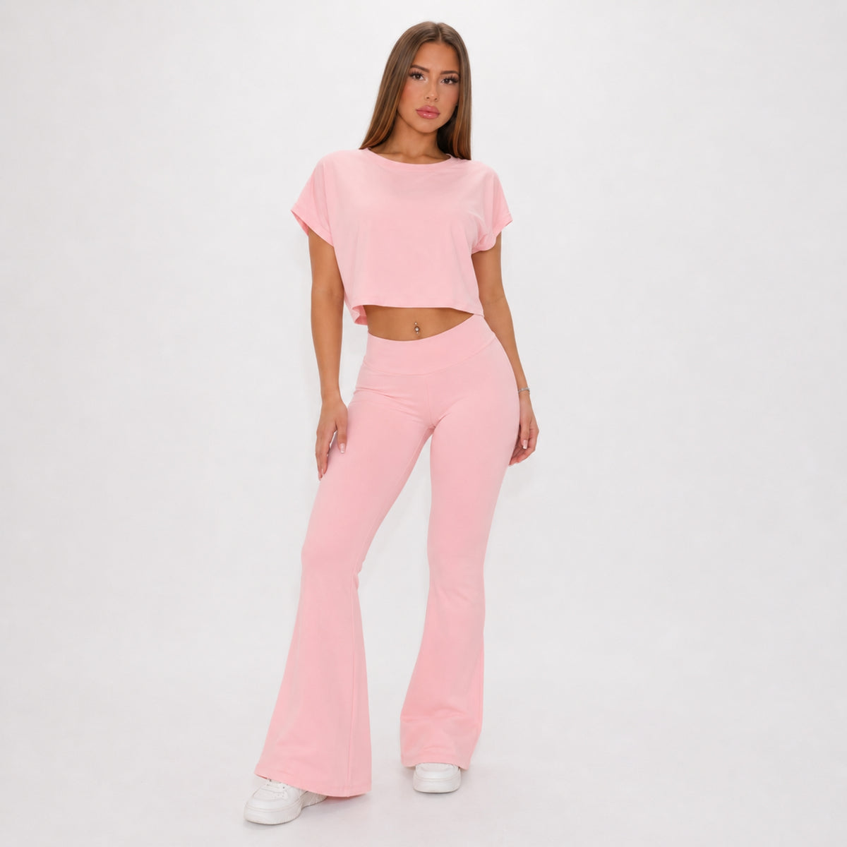 A woman modeling a light pink two-piece outfit consisting of a short-sleeve cropped t-shirt and matching high-waisted flare pants, paired with white sneakers against a plain white studio background.