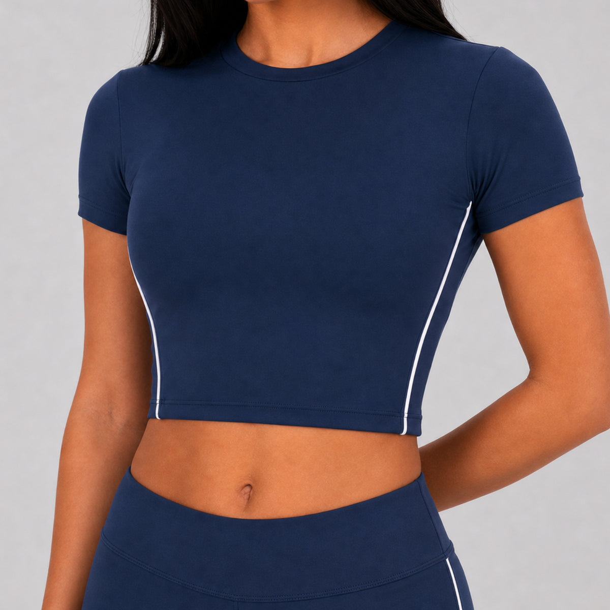 A close-up of a model wearing a navy blue short-sleeve crop top with white piping details along the sides, paired with matching navy athletic bottoms.