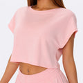 A woman wearing a light pink short-sleeve crop top and matching pink pants, photographed against a plain light gray studio background.