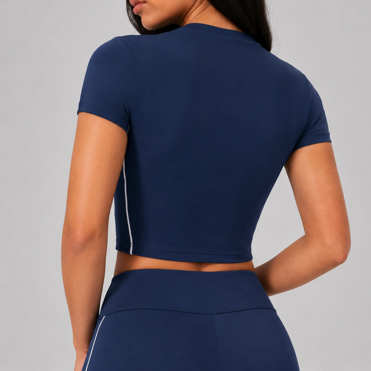 A woman shown from the back wearing a navy blue short-sleeve crop top with a thin white side stripe and matching navy bottoms, against a plain light gray studio background.