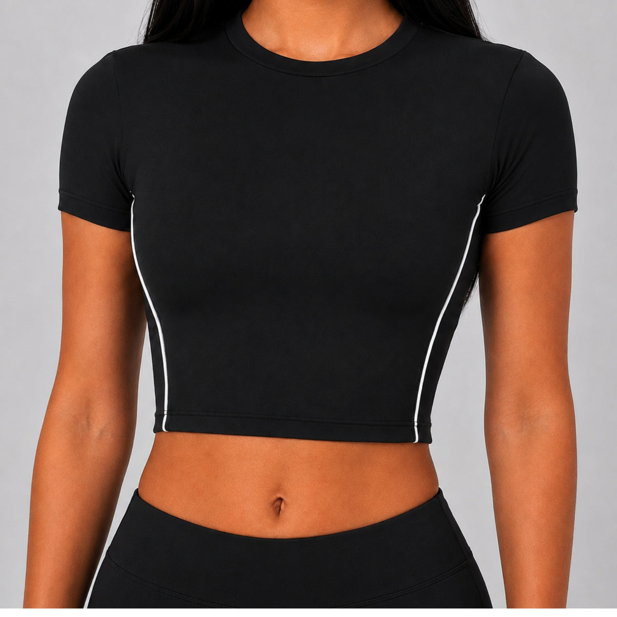 A woman wearing a fitted black short-sleeve crop top with thin white side piping, shown from the neck to the hips against a light gray studio background.