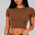 A woman wearing a fitted brown short-sleeve crop top with thin white piping along the sides, shown against a light gray studio background.