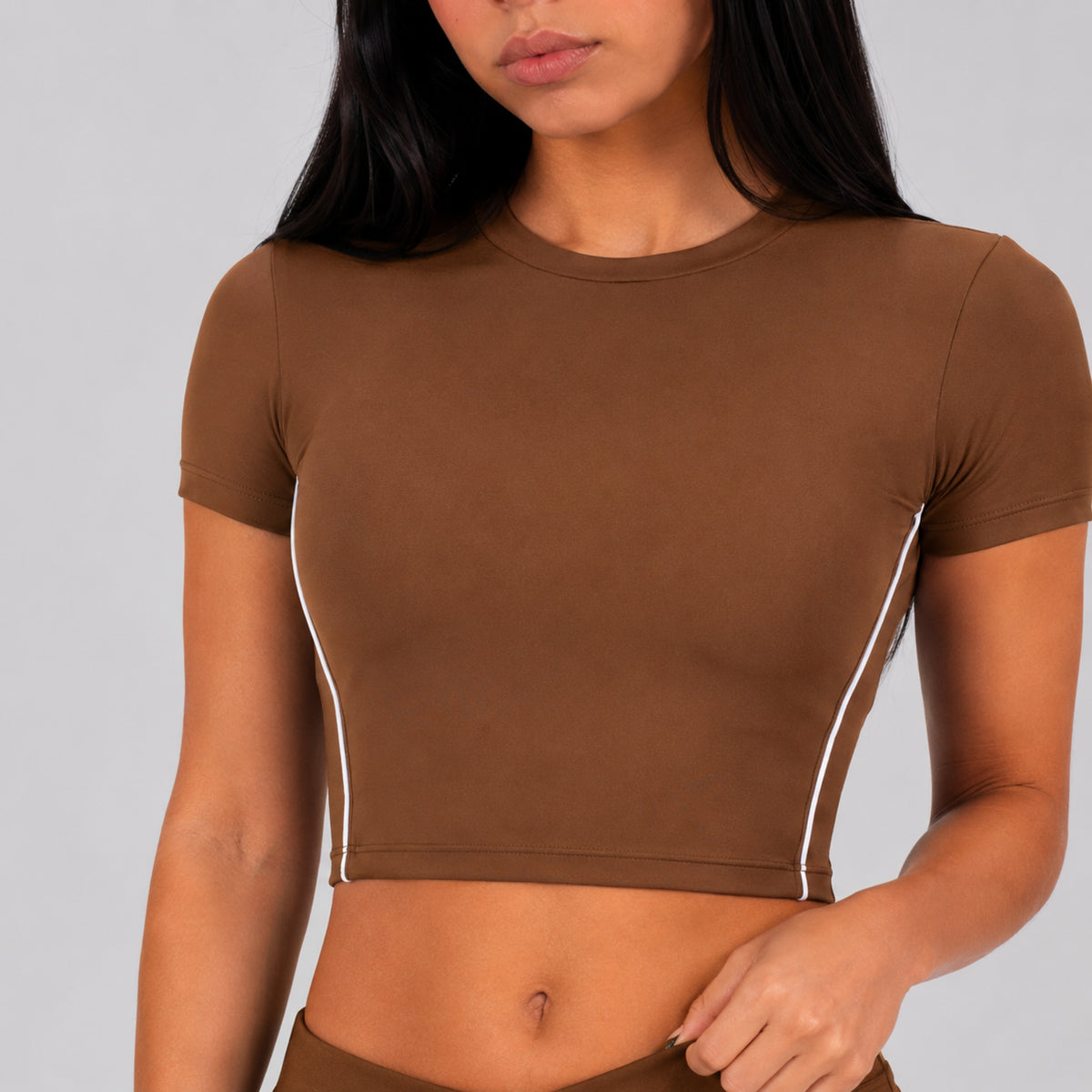 A woman wearing a fitted brown short-sleeve crop top with thin white piping along the sides, shown against a light gray studio background.