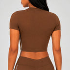 Rear view of a woman wearing a brown short-sleeve crop top and matching brown bottoms against a light gray studio background.