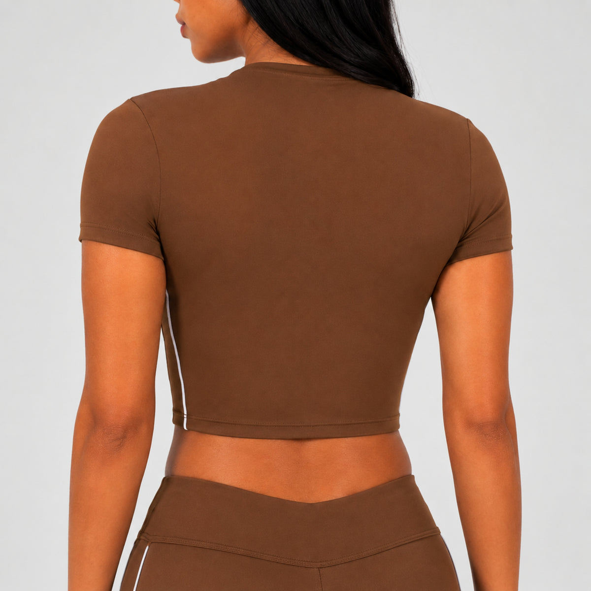 Rear view of a woman wearing a brown short-sleeve crop top and matching brown bottoms against a light gray studio background.