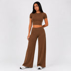 A woman modeling a matching brown two-piece set consisting of a short-sleeve crop top and high-waisted wide-leg pants, photographed against a plain light gray studio background.