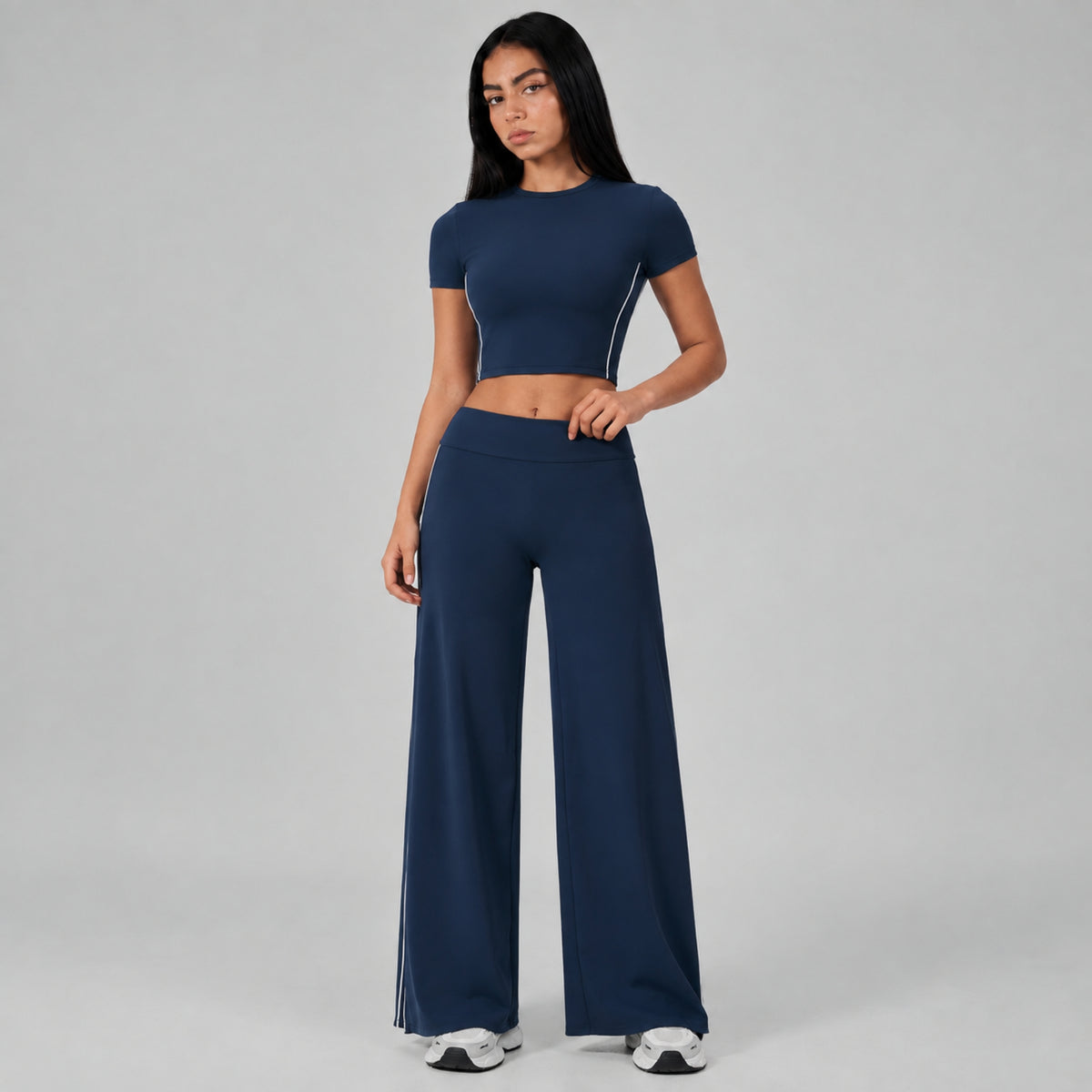 A woman modeling a matching navy blue activewear set consisting of a short-sleeve crop top with white side piping and high-waisted wide-leg pants with white side piping, set against a plain gray studio background.
