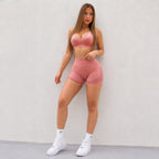 A woman modeling a matching pink activewear set consisting of a sports bra and high-waisted shorts, paired with white socks and white sneakers, posing against a light gray textured wall.