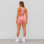 A woman is shown from behind wearing a matching pink athletic set consisting of a sports bra and high-waisted shorts with a textured pattern, paired with white sneakers, against a plain light gray wall.