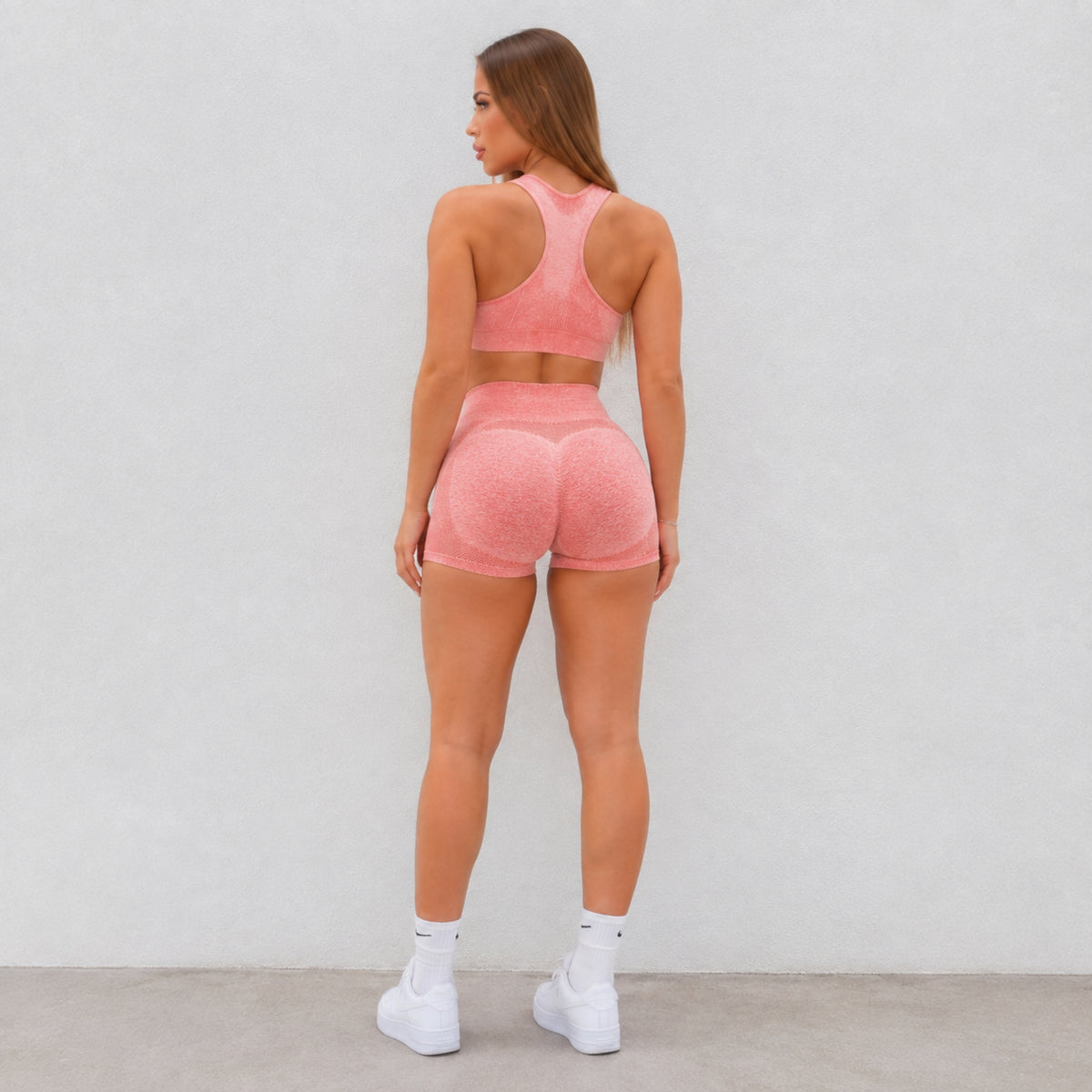 A woman is shown from behind wearing a matching pink athletic set consisting of a sports bra and high-waisted shorts with a textured pattern, paired with white sneakers, against a plain light gray wall.
