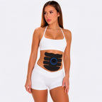 A woman modeling a white sports bra and matching shorts with a black abdominal muscle stimulator pack applied to her stomach, set against a plain light gray studio background.