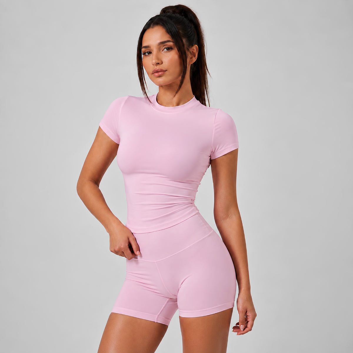 A woman modeling a light pink short-sleeve athletic bodysuit with matching high-waisted shorts against a solid light gray studio background.