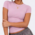 A close-up of a woman wearing a fitted pink short-sleeve athletic top with a high crew neck and ruched waist details, paired with matching leggings, against a clean background.