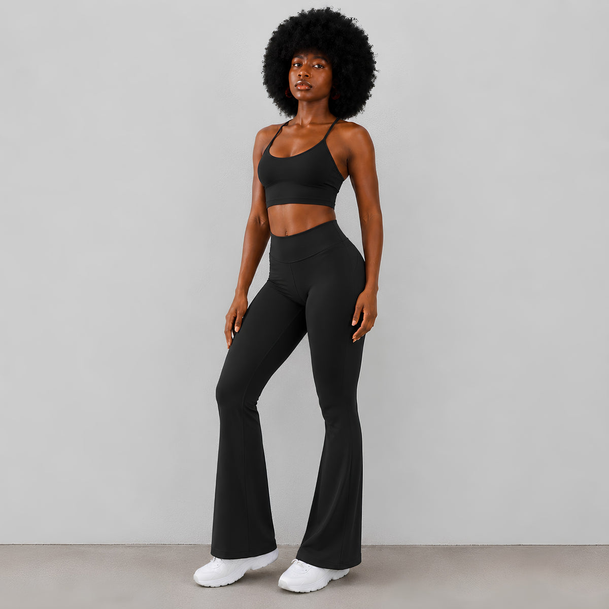 A woman modeling a black two-piece activewear set consisting of a sports bra and high-waisted flared leggings, paired with sneakers, against a plain studio background.