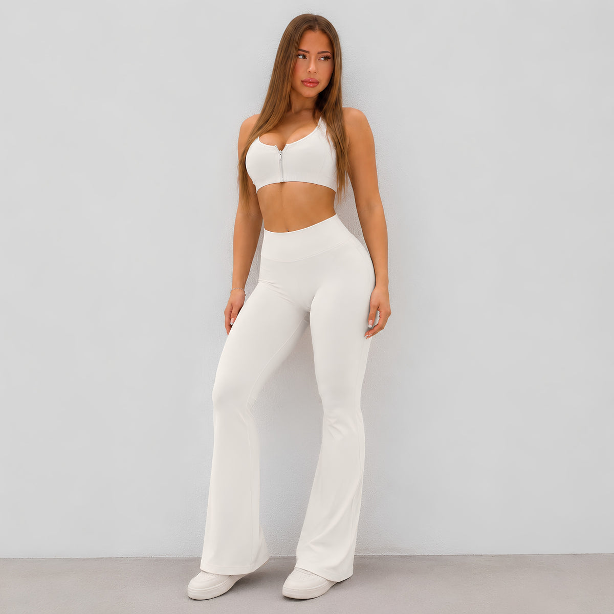 A woman modeling a white two-piece activewear set consisting of a halter crop top and high-waisted flared pants, standing against a light gray studio wall.
