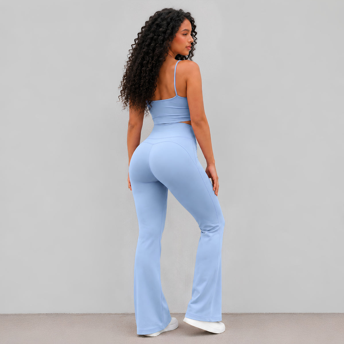 A woman with long curly dark hair is shown from the back wearing a light blue high-waisted activewear set consisting of a spaghetti-strap crop top and matching flared leggings, paired with sneakers against a plain light studio background.