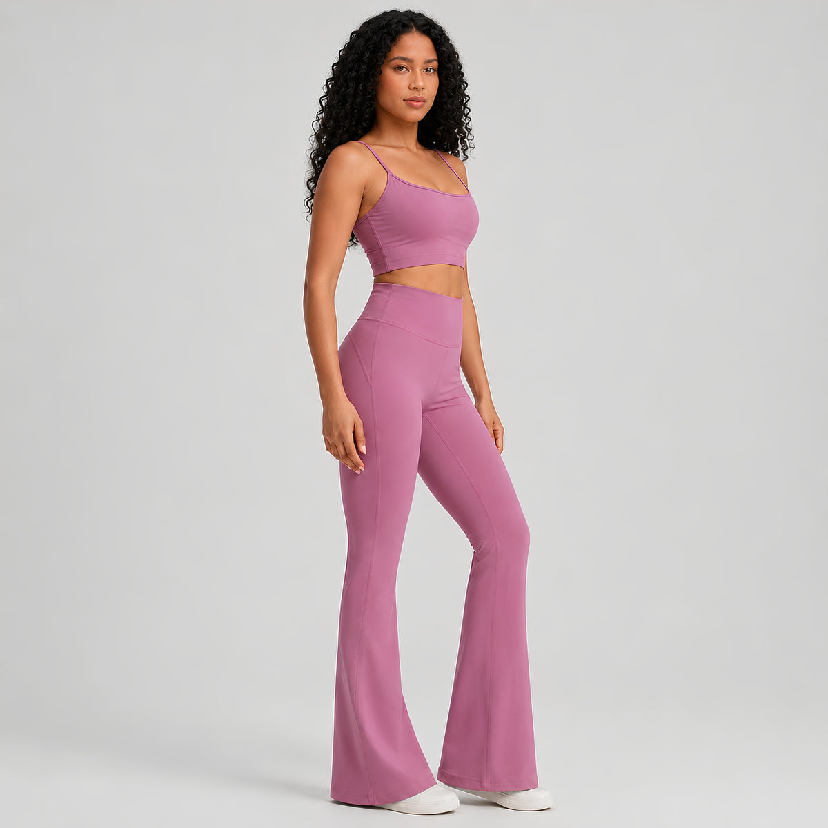 A woman modeling a matching dusty pink activewear set consisting of a spaghetti-strap crop top and high-waisted flared leggings, set against a plain light studio background.