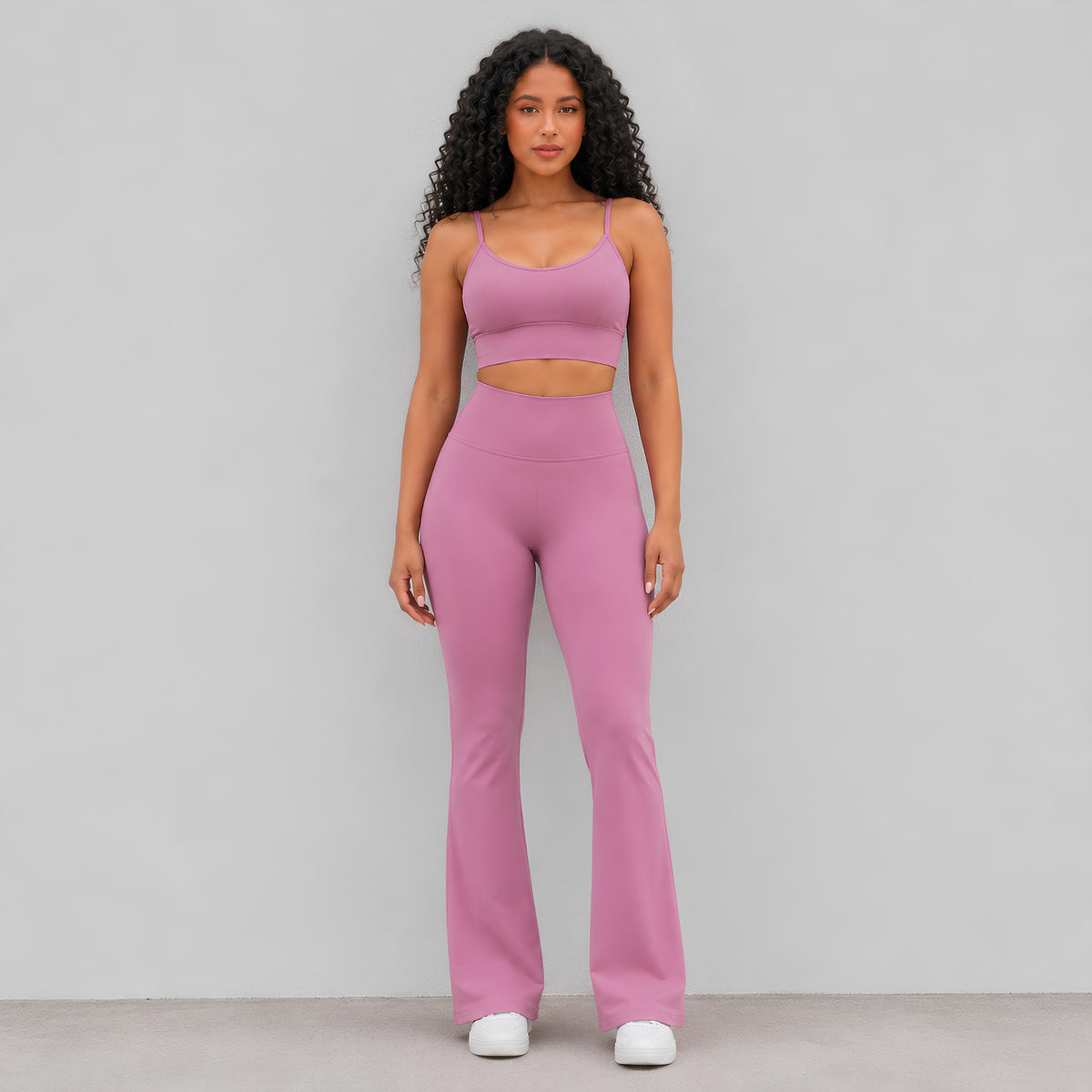 A woman modeling a matching dusty pink activewear set consisting of a sports bra and high-waisted flared leggings, standing against a plain light wall.