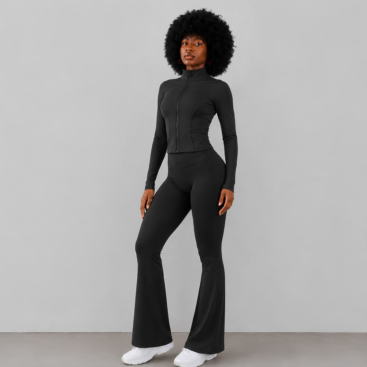 A model with an afro hairstyle wearing a black long-sleeve mock-neck zip-up jumpsuit with flared pants and sneakers, standing against a light background.