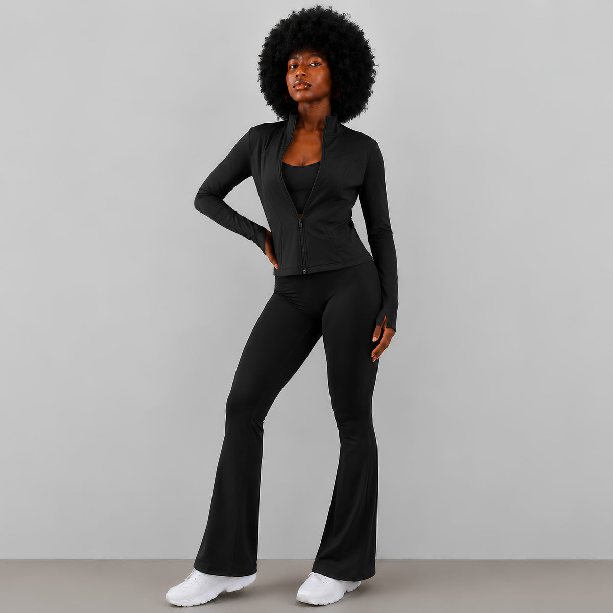 A woman modeling a black two-piece activewear set consisting of a long-sleeve zip-up jacket and matching flared pants, paired with sneakers, against a solid studio background.
