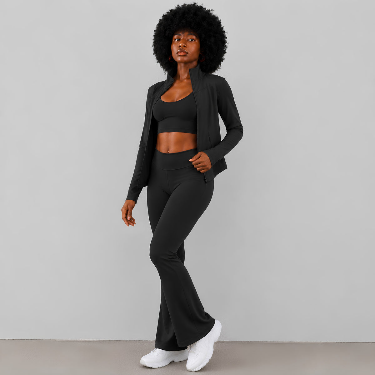 A woman modeling a black activewear set consisting of a cropped sports bra, a matching open-front cardigan, high-waisted flared leggings, and sneakers, set against a plain studio background.