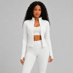 A woman modeling a white athletic set consisting of a cropped zip-up jacket, sports bra, and leggings against a solid studio background.