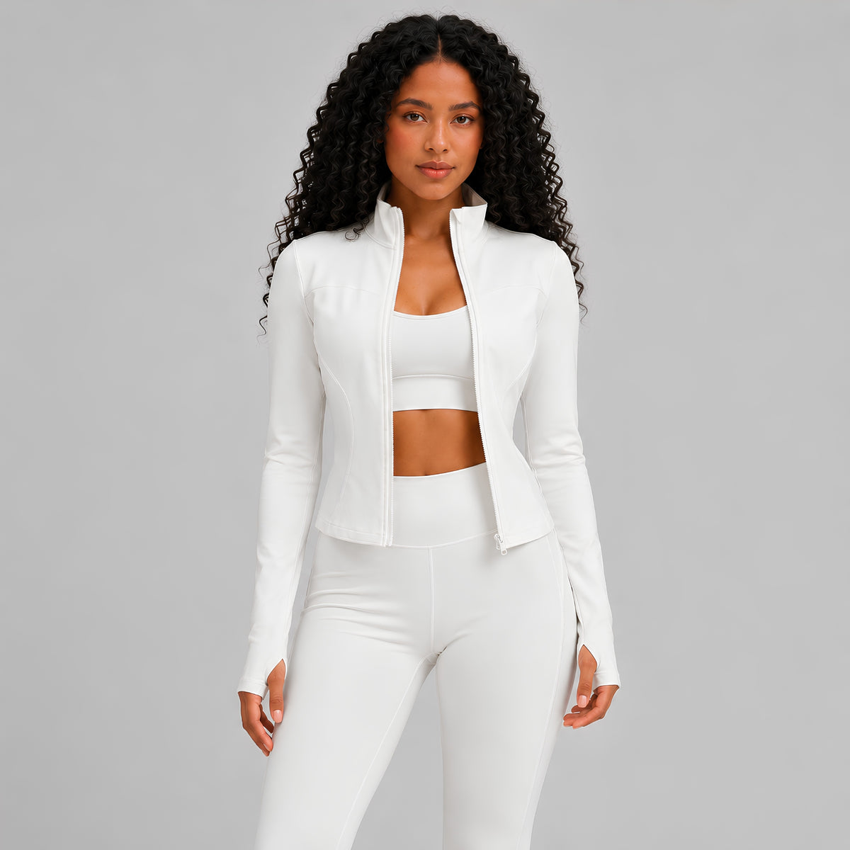 A woman modeling a white athletic set consisting of a cropped zip-up jacket, sports bra, and leggings against a solid studio background.