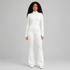 A model with long curly hair wearing a matching white long-sleeve zip-up top and white flared pants, standing against a light studio background.