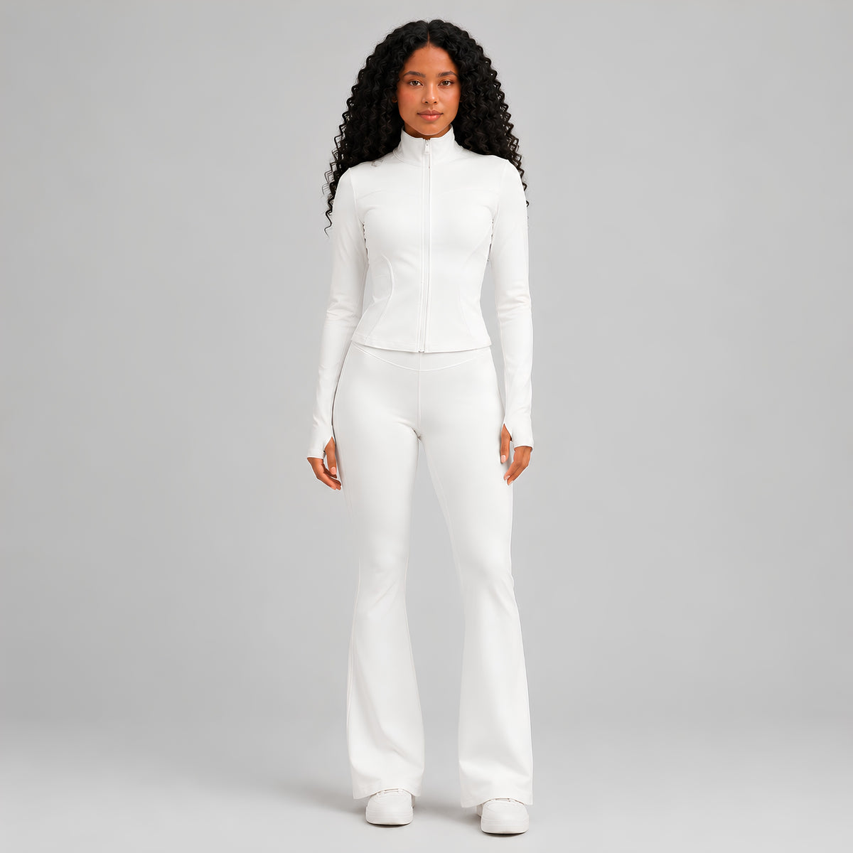 A model with long curly hair wearing a matching white long-sleeve zip-up top and white flared pants, standing against a light studio background.