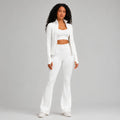 A woman modeling a matching white activewear set consisting of a long-sleeve cropped blazer, a scoop-neck crop top, high-waisted flared leggings, and white sneakers, shot against a solid light studio background.
