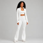 A woman modeling a matching white activewear set consisting of a long-sleeve cropped blazer, a scoop-neck crop top, high-waisted flared leggings, and white sneakers, shot against a solid light studio background.