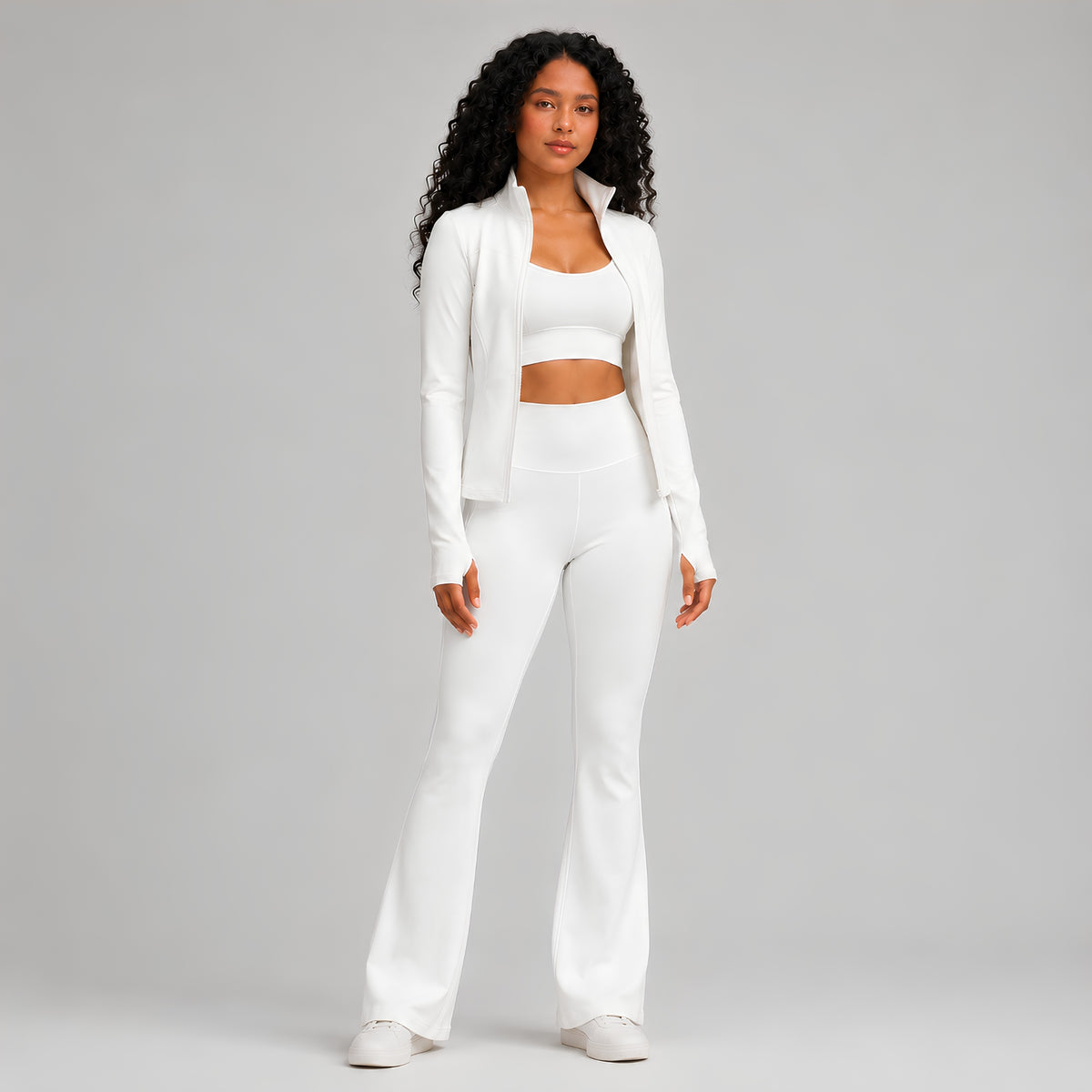 A woman modeling a matching white activewear set consisting of a long-sleeve cropped blazer, a scoop-neck crop top, high-waisted flared leggings, and white sneakers, shot against a solid light studio background.