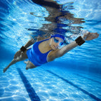 A woman underwater in a swimming pool wearing a blue one-piece swimsuit, blue swim goggles, and black wristbands.